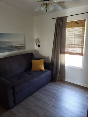 A room with a dark blue sofa, a single mustard-colored pillow, a ceiling fan, a wall-mounted lamp, and a window with blinds. - Bayfront cottage offers amazing views of the water and Assateague Island (Berlin)