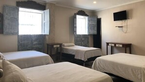 Habitación de hotel con tres camas individuales, una mesita, televisor montado en la pared y ventana con cortinas.