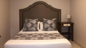Un dormitorio con una cama grande, cabecera de madera, dos almohadas, una mesita de noche con lámpara y un equipo de aire acondicionado en la pared.