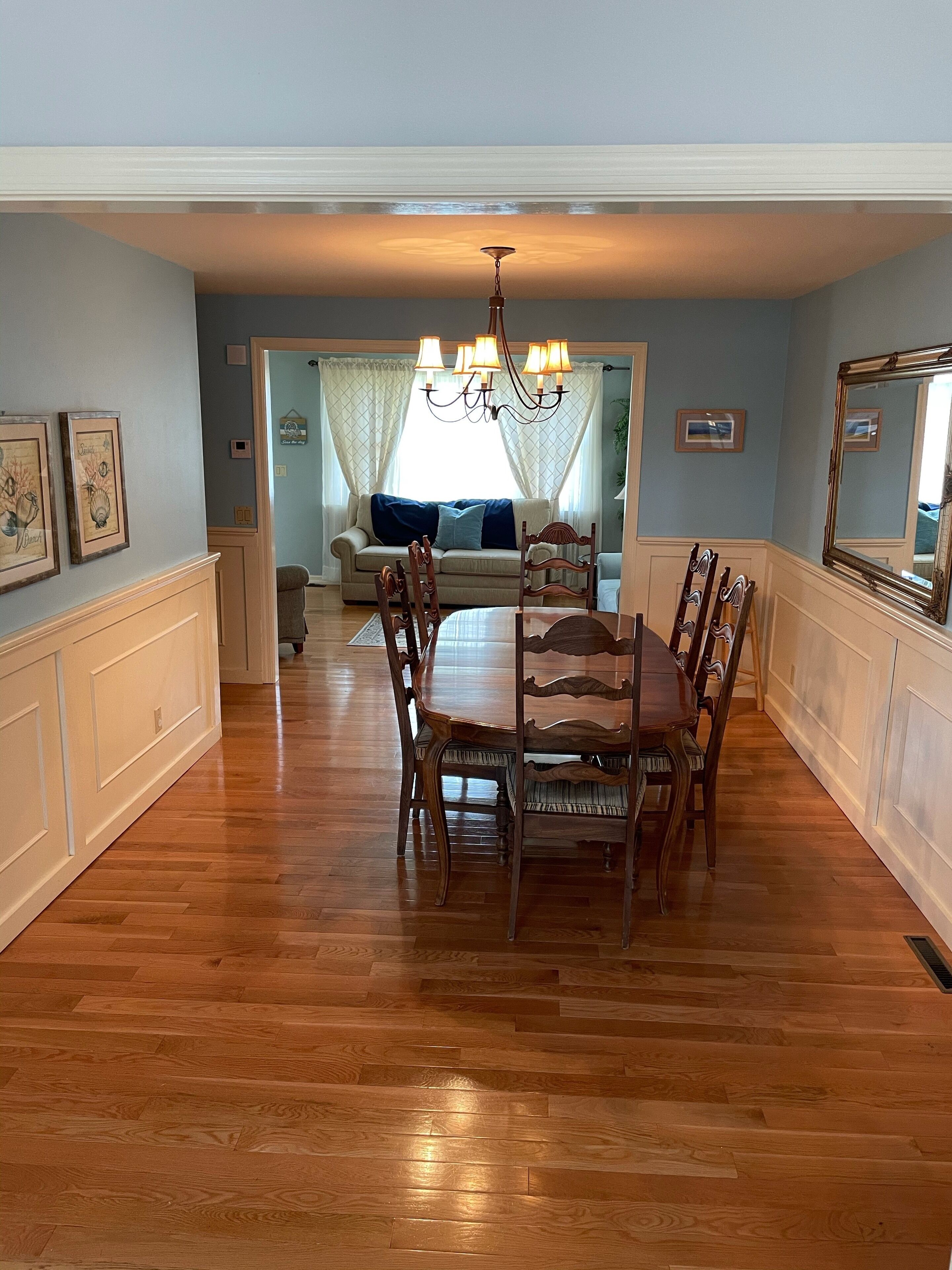A dining area with a wooden table and chairs, a chandelier, and a view into a living room with a sofa and curtains.