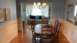 A dining area with a wooden table and chairs, a chandelier, and a view into a living room with a sofa and curtains. - Perfect Family/Friends Reunion House in the Hamptons! Close to Ocean Beaches! (Hampton Bays)