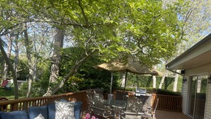 A covered outdoor patio with a wooden deck, a glass-top table, a sofa with patterned cushions, and a grill. - Perfect Family/Friends Reunion House in the Hamptons! Close to Ocean Beaches! (Hampton Bays)