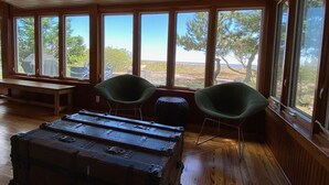 Ein Zimmer mit großen Fenstern, einer hölzernen Bank, zwei grünen Stühlen und einem Vintage-Koffer.
