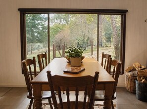A dining area with a large table, wooden chairs, a potted plant, and a view of a garden through large windows.