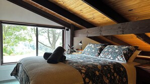 A bedroom with a large window, wooden ceiling beams, a bed with floral bedding, and a bedside lamp. - Top of the Otways - Nature Lodge (Barramunga)