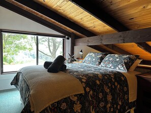 A bedroom with a large window, wooden ceiling beams, a bed with floral bedding, and a bedside lamp. - Top of the Otways - The Nature Lodge (Barramunga)