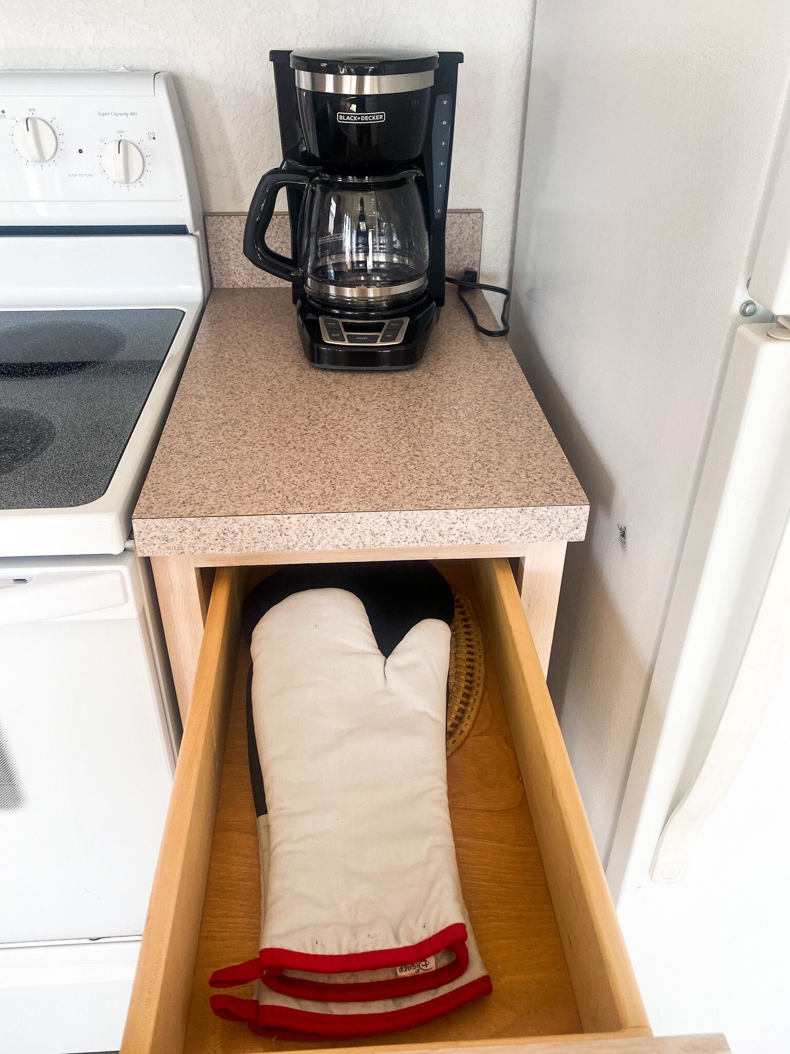Une petite cuisine équipée d’une machine à café, d’une cuisinière et d’un tiroir contenant un manique de couleur blanche.