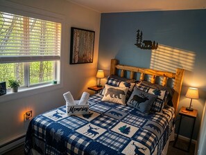 Ein Schlafzimmer mit kariertem Bettbezug, einem Kopfteil, Nachttischlampen, einem Wandbild und einem Fenster mit Jalousien.