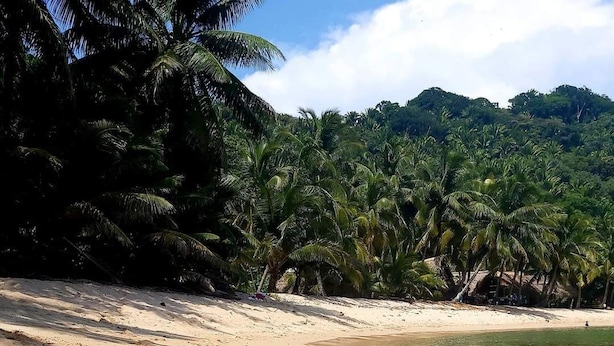Une personne allongée sur une plage de sable fin, avec une eau cristalline, entourée de palmiers et avec une chaîne de montagnes en arrière-plan.