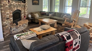 Un salon chaleureux avec une cheminée en briques, un canapé d’angle, une table basse en bois et une grande fenêtre laissant entrer la lumière naturelle.