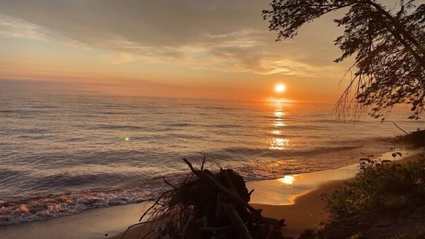 Une scène de plage avec un coucher de soleil sur l’océan, des vagues calmes et un tas de bois flotté sur le rivage.