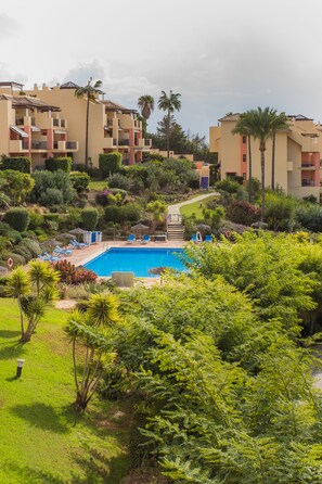 Un área de piscina con sillones de descanso y una piscina azul cristalina rodeada de exuberante vegetación y edificios de varios pisos.
