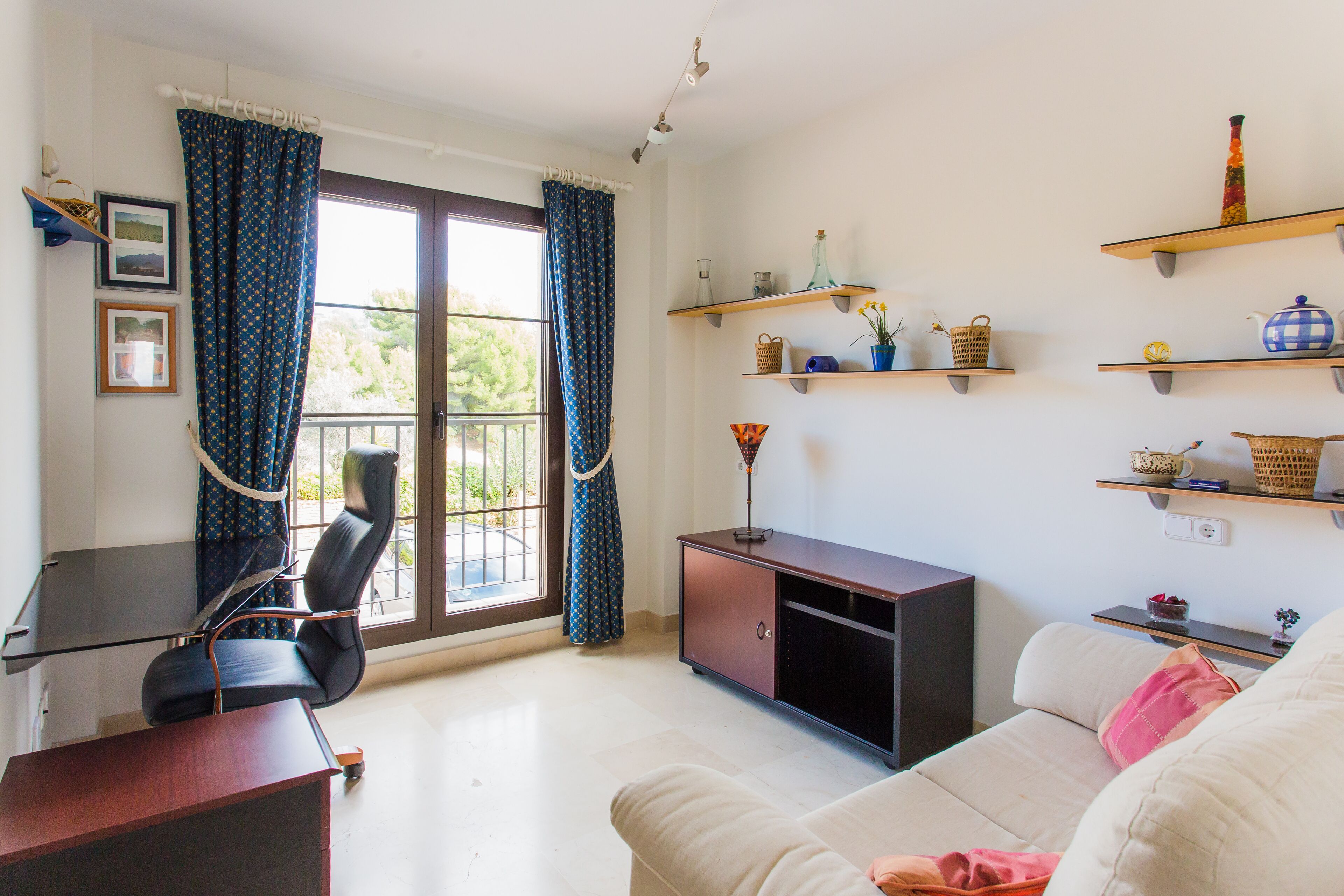 A living room with a sofa, a desk, and a chair. There are shelves with decorative items and a window with curtains.