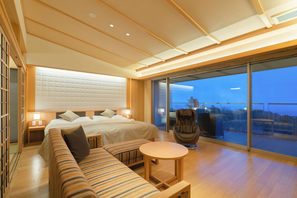 A modern hotel room with a large bed, a sofa, a small table, and a balcony with a view.