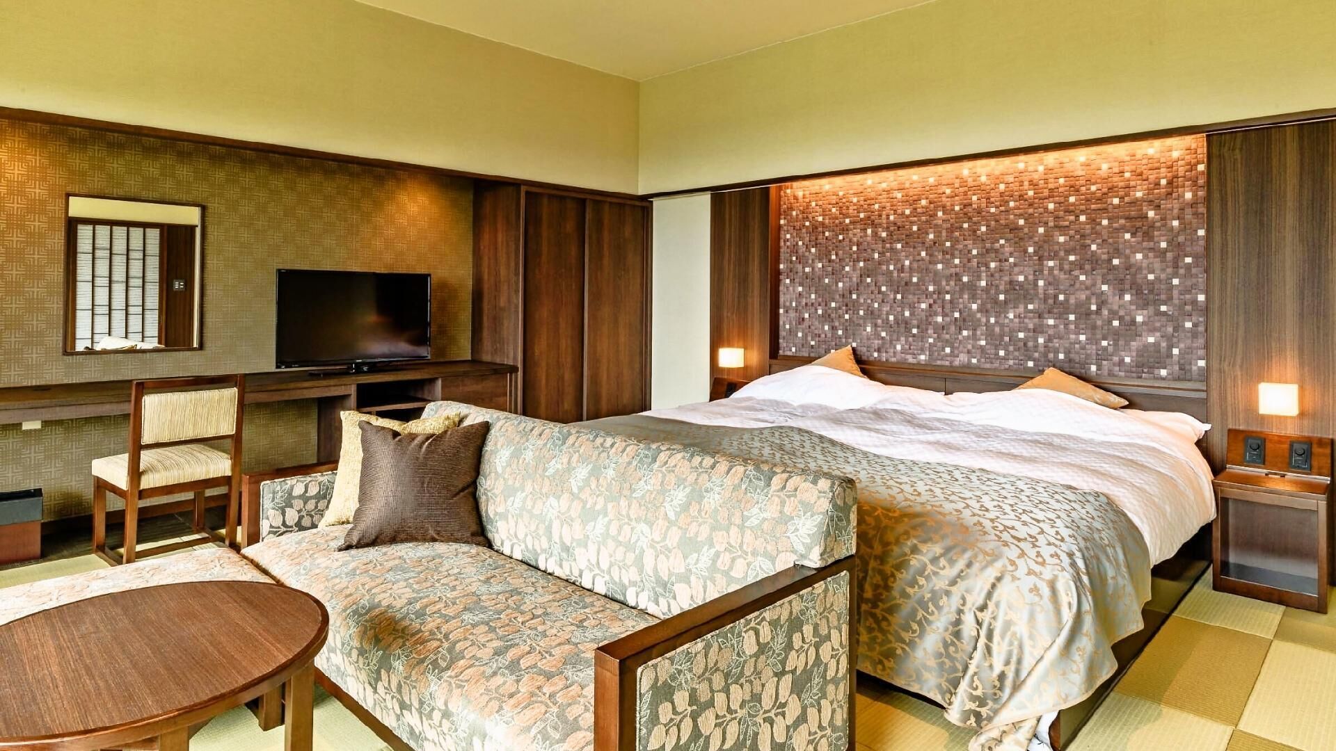 A hotel room with a bed, a sofa, a TV, a desk, and a wall-mounted light.
