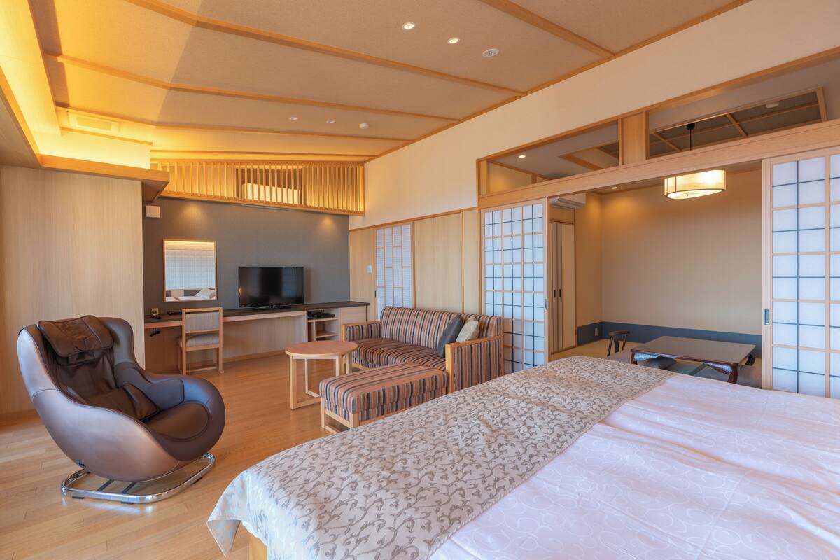 A hotel room with a bed, a massage chair, a desk with a TV, and a seating area with a table and chairs.