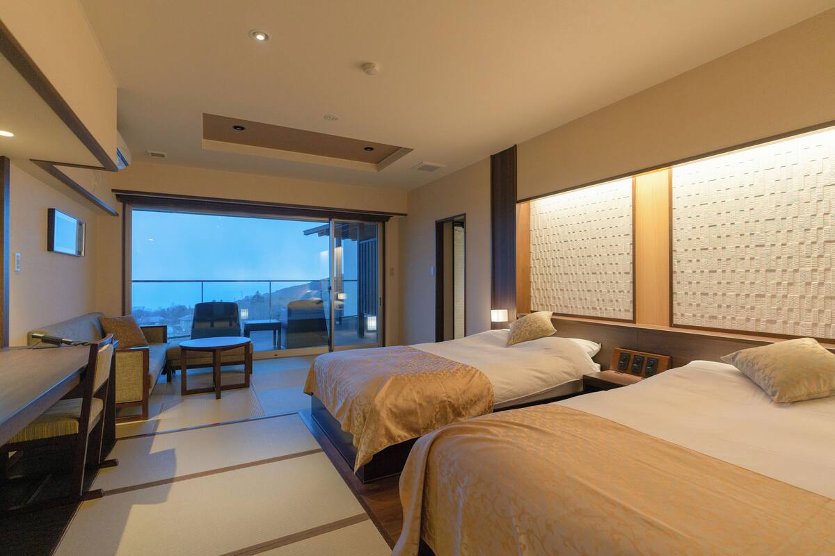 A hotel room with two beds, a sofa, a desk, and a view of the outdoors.