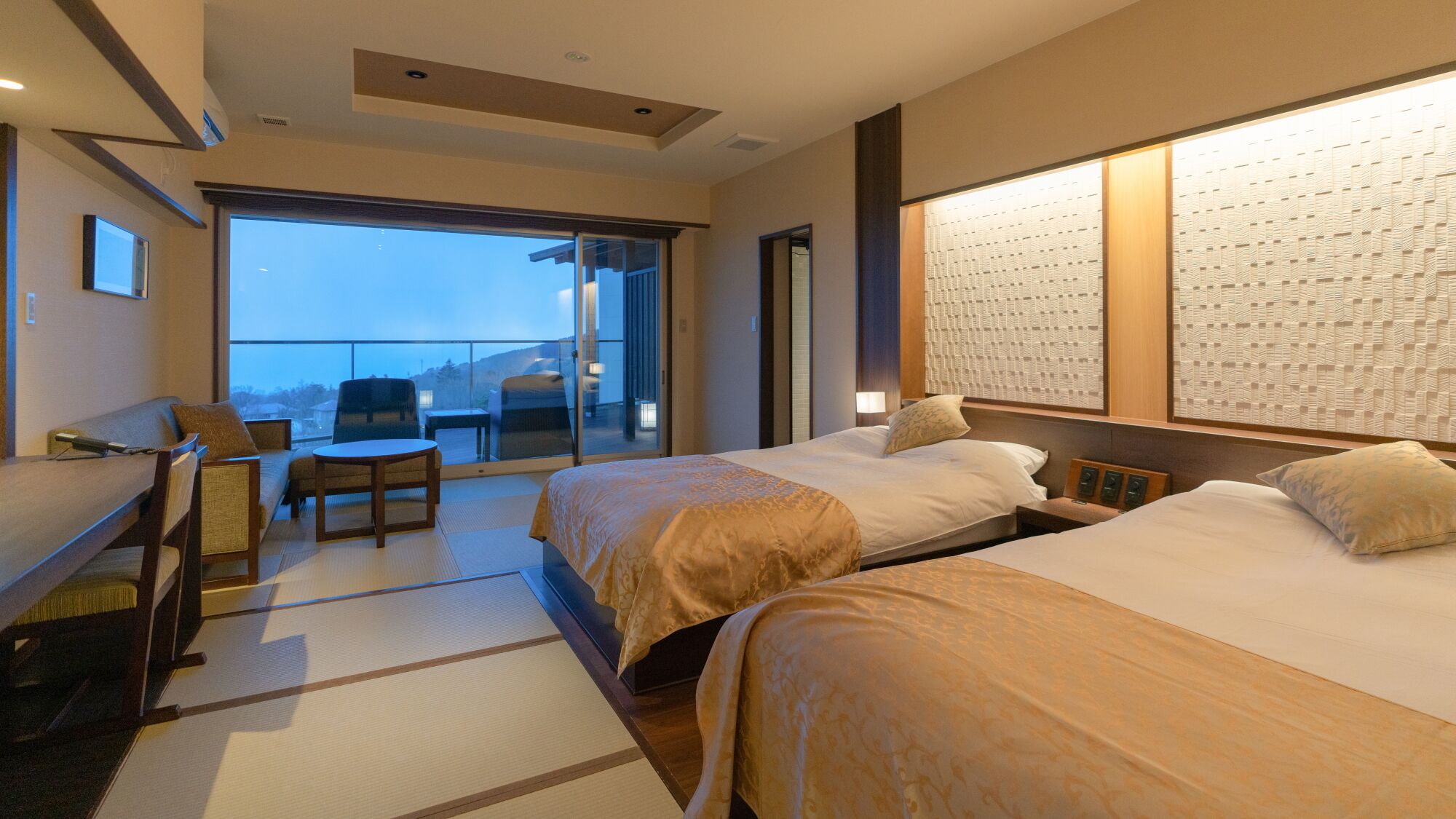 A hotel room with two beds, a sofa, a small table, and a balcony with a view.