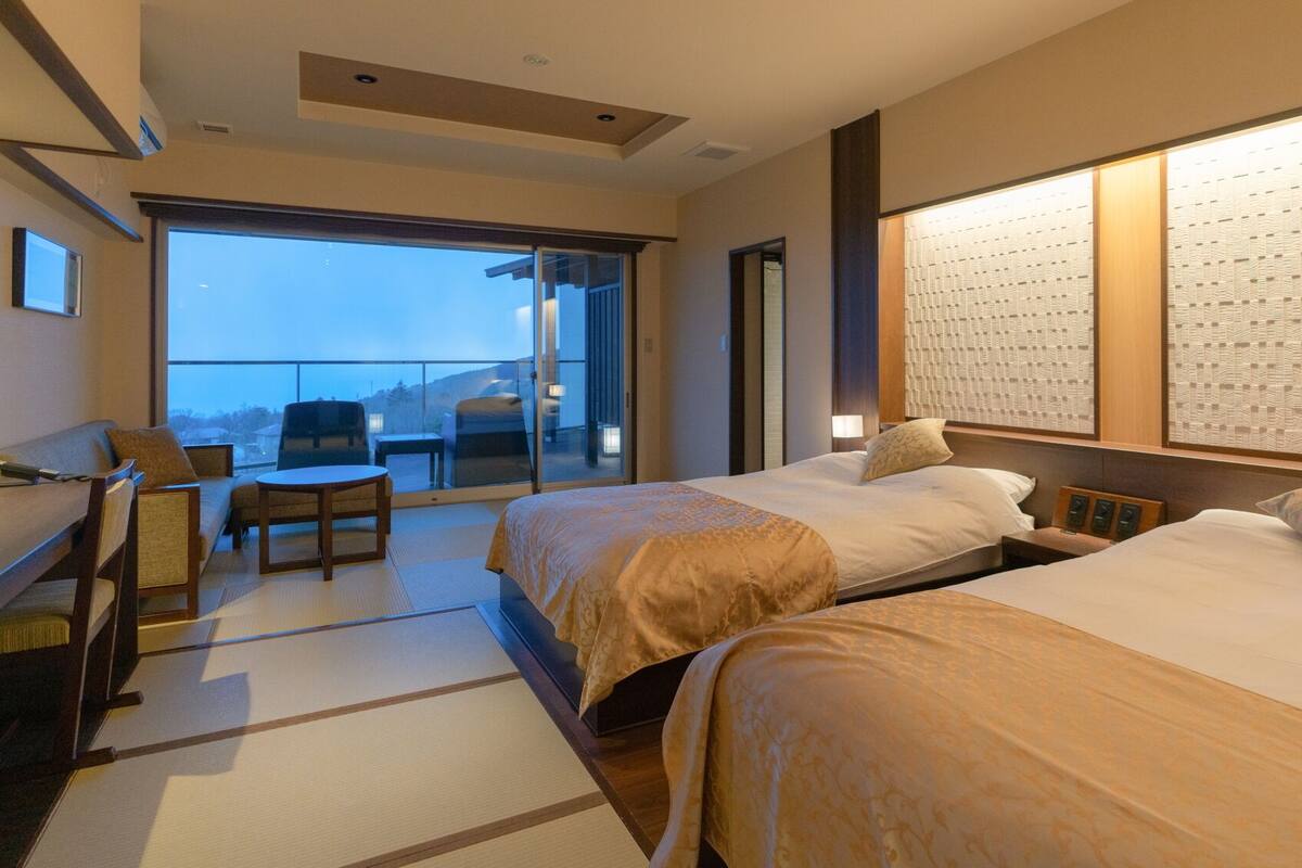 A hotel room with two beds, a sofa, a small table, and a balcony with a view.