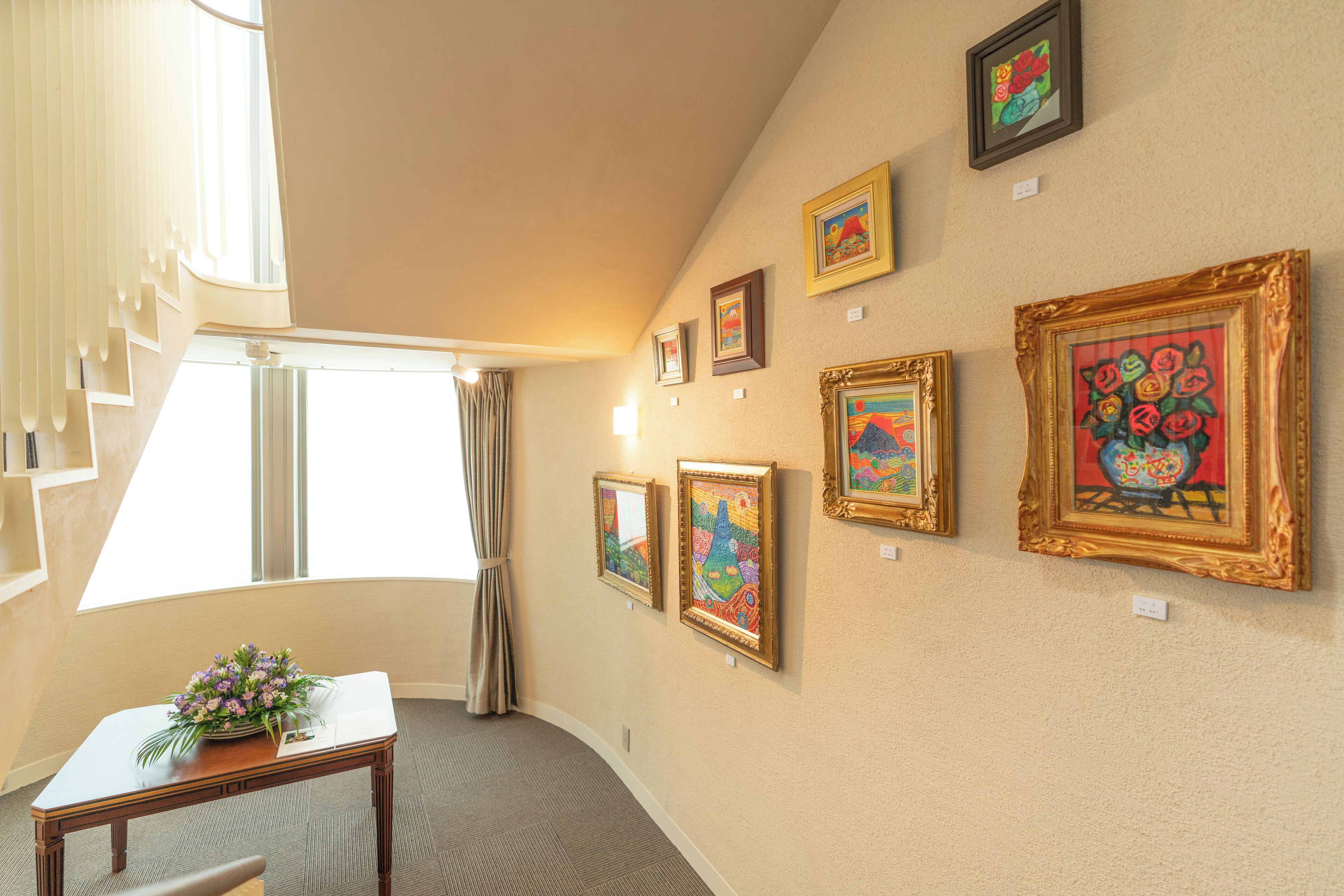 A hotel room with a curved ceiling, a window with curtains, a table with a vase of flowers, and several framed artworks on the wall.