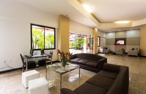 A modern living room with a brown sofa, a glass coffee table, and a desk with a computer. There is a TV mounted on the wall and a window with a view of greenery outside. - Joh Blue Ocean Hotel (Fortaleza)