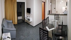 A compact living space with a grey sofa, a dining table with chairs, a TV mounted on a white wall, and a kitchen area with a microwave. - JOH Blue Ocean Hotel (Fortaleza)