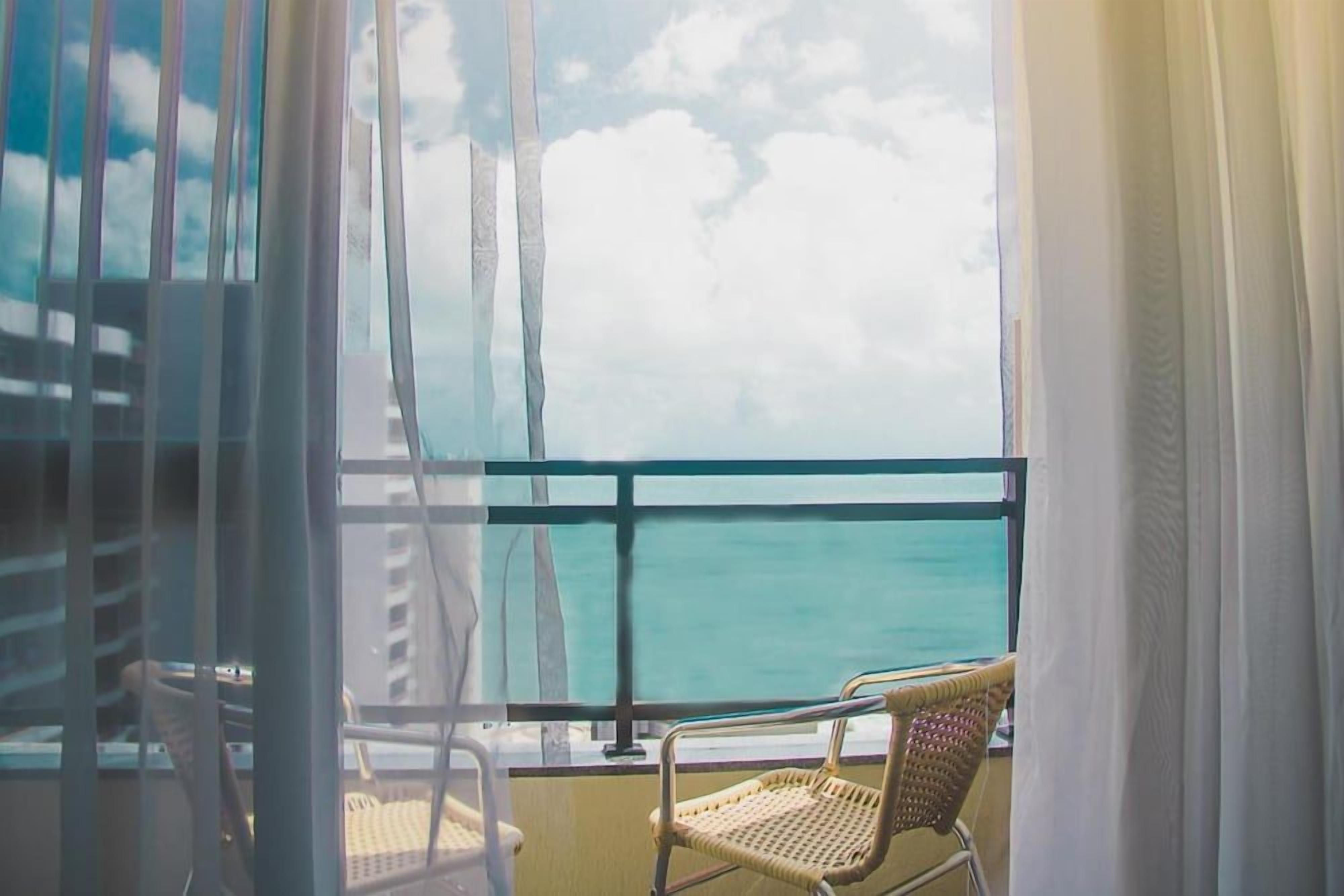 A balcony with a view of the sea, a wicker chair, and a window with curtains.