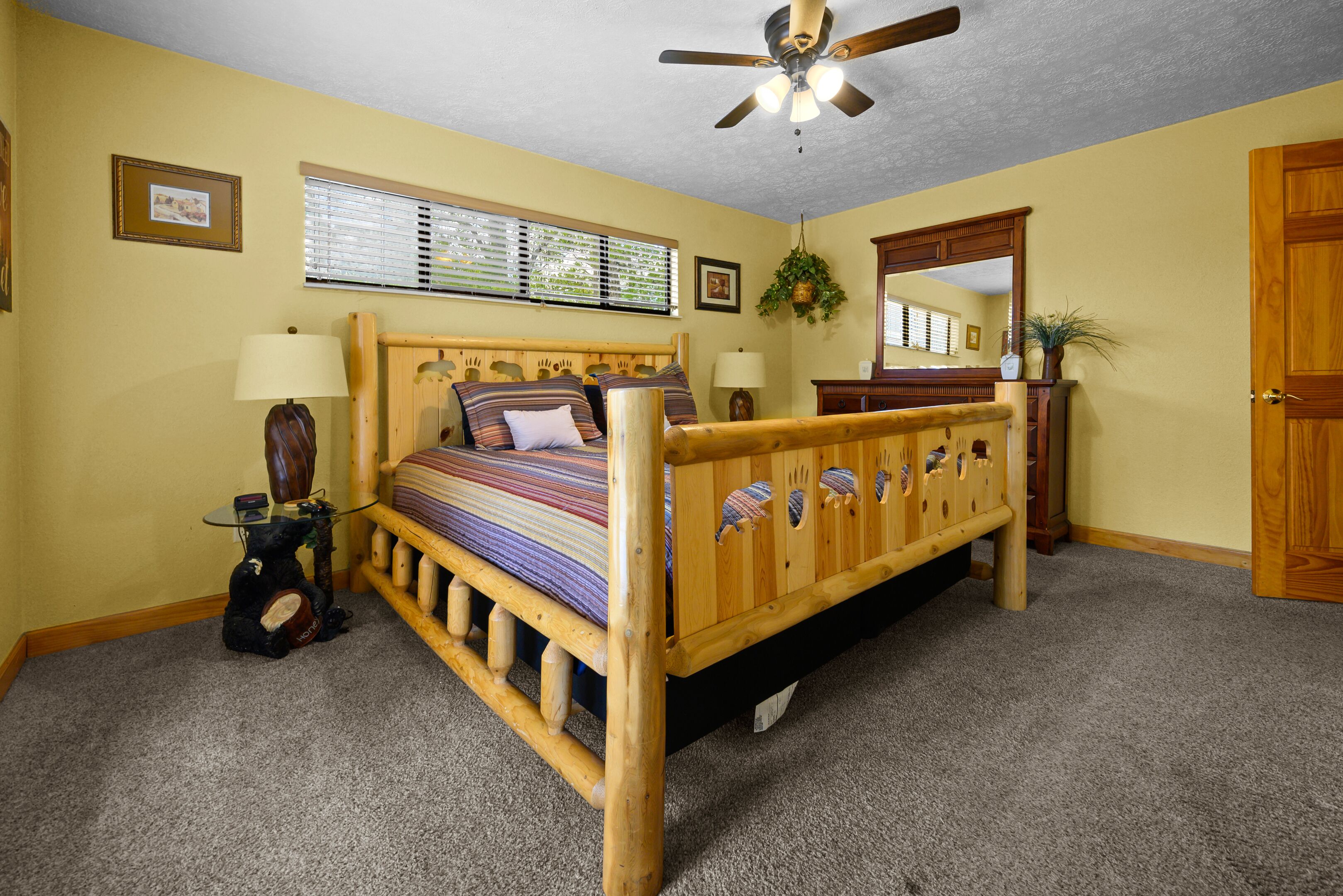A bedroom with a wooden bed, a ceiling fan, a window with blinds, a lamp, a guitar, and a mirror.