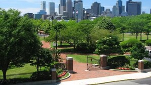 A city skyline with modern high-rise buildings, a well-maintained park with trees and a paved walkway, and a gated entrance. - Beautiful 4BD/2BA Gem! Near airport, city, subway. (Boston)