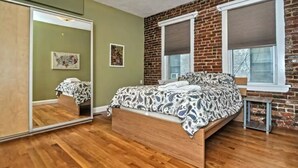 A bedroom with a wooden floor, a bed with a patterned bedspread, a large mirror, and a brick wall. - Beautiful 4BD/2BA Gem! Near airport, city, subway. (Boston)