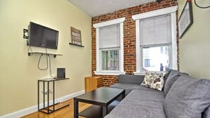 A living room with a gray sofa, a black coffee table, a TV on a stand, and a brick wall. - Beautiful 4BD/2BA Gem! Near airport, city, subway. (Boston)