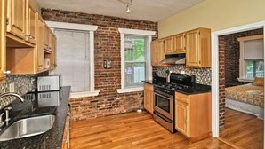 A compact kitchen with wooden cabinets, a black countertop, and a brick wall. - Beautiful 4BD/2BA Gem! Near airport, city, subway. (Boston)