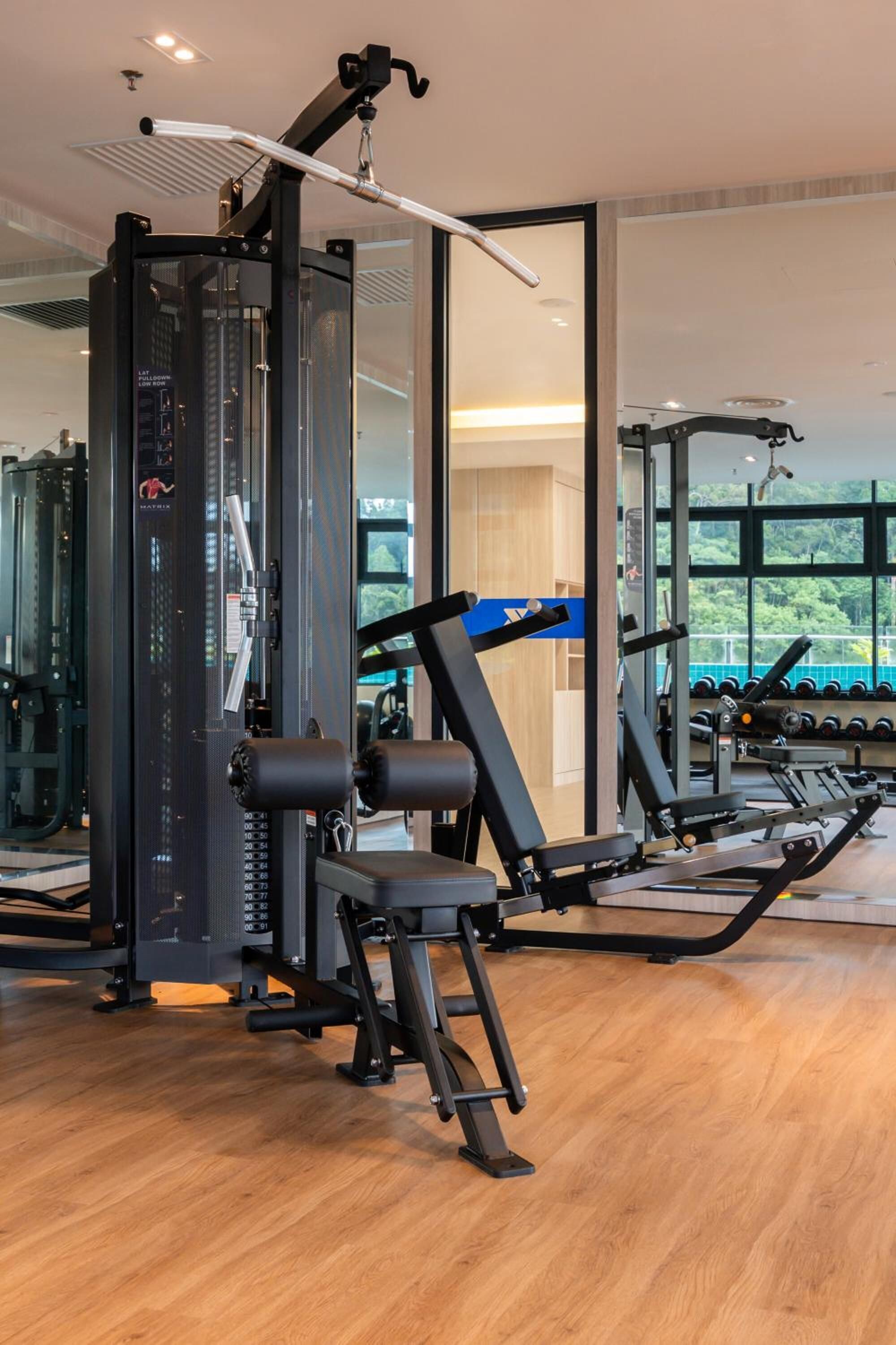 Un gimnasio con diferentes máquinas de ejercicio, un espejo grande y vistas al exterior con vegetación.