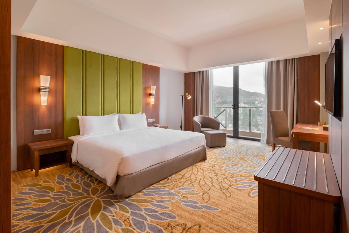 A hotel room with a large bed, a desk, a chair, a TV, and a balcony with a view.