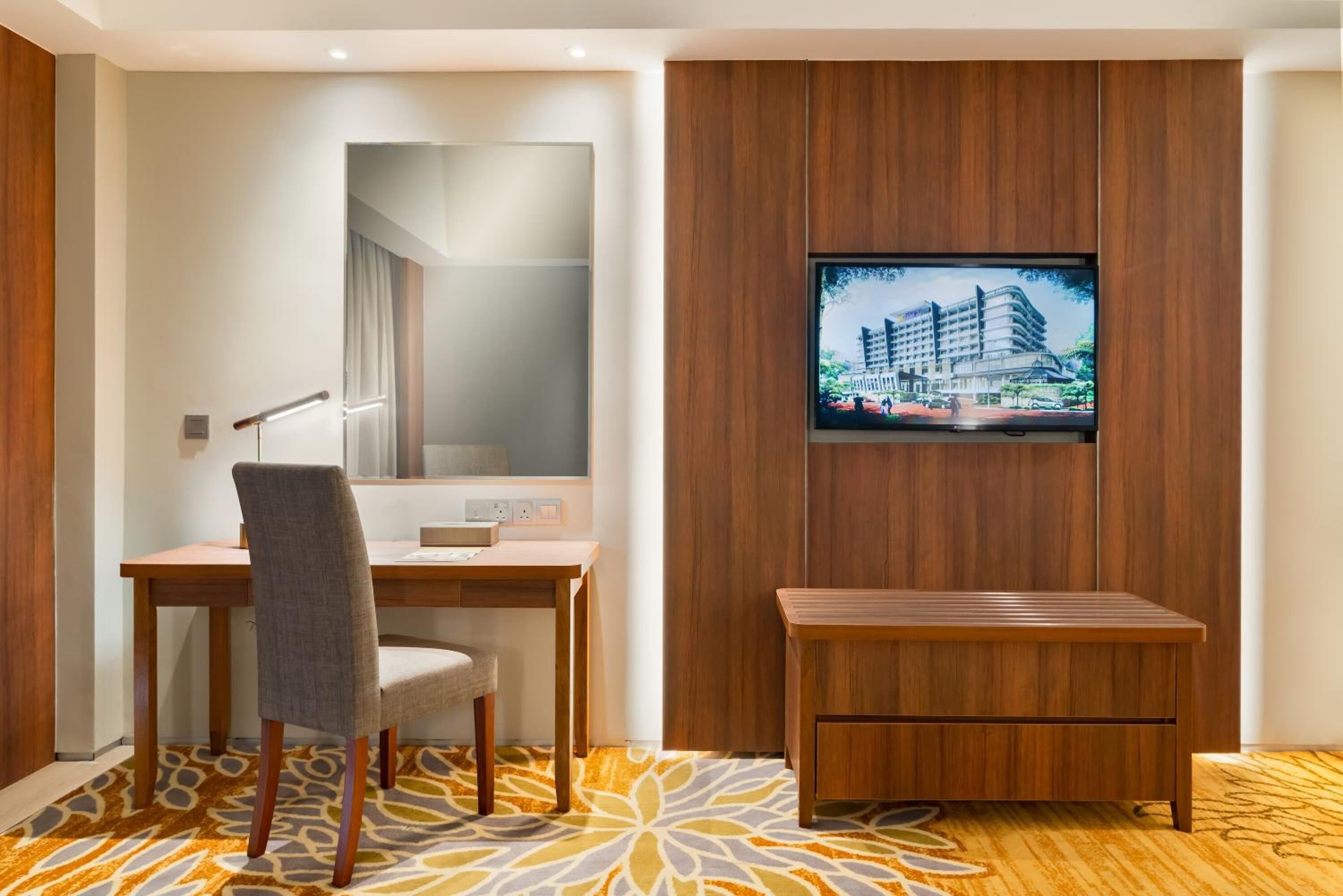 A hotel room with a wooden desk, a chair, a television mounted on the wall, and a patterned carpet.