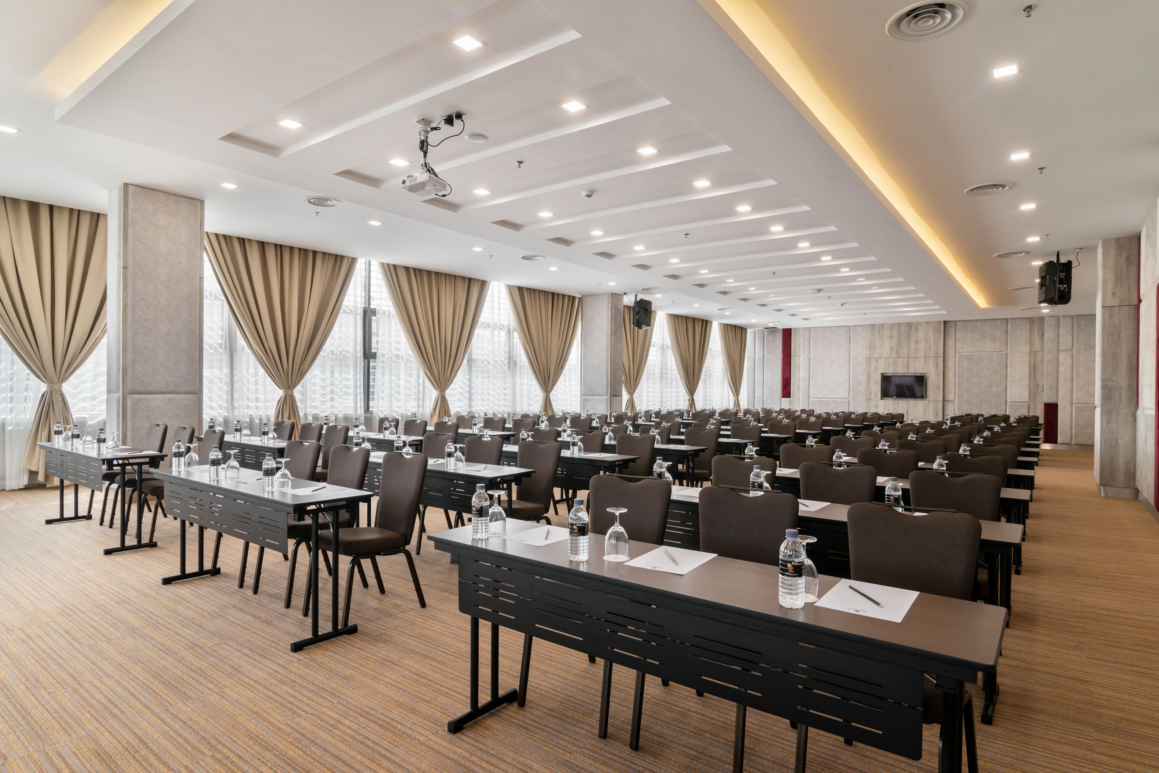 Un salón de conferencias con filas de mesas y sillas, ventanas amplias con cortinas y un techo con iluminación empotrada.