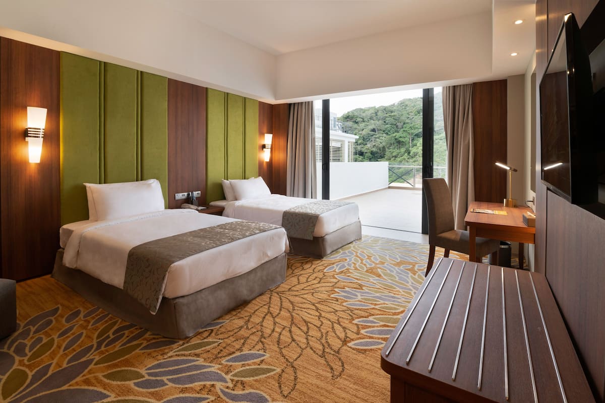 A hotel room with two beds, a desk, a TV, and a balcony with a view of greenery.