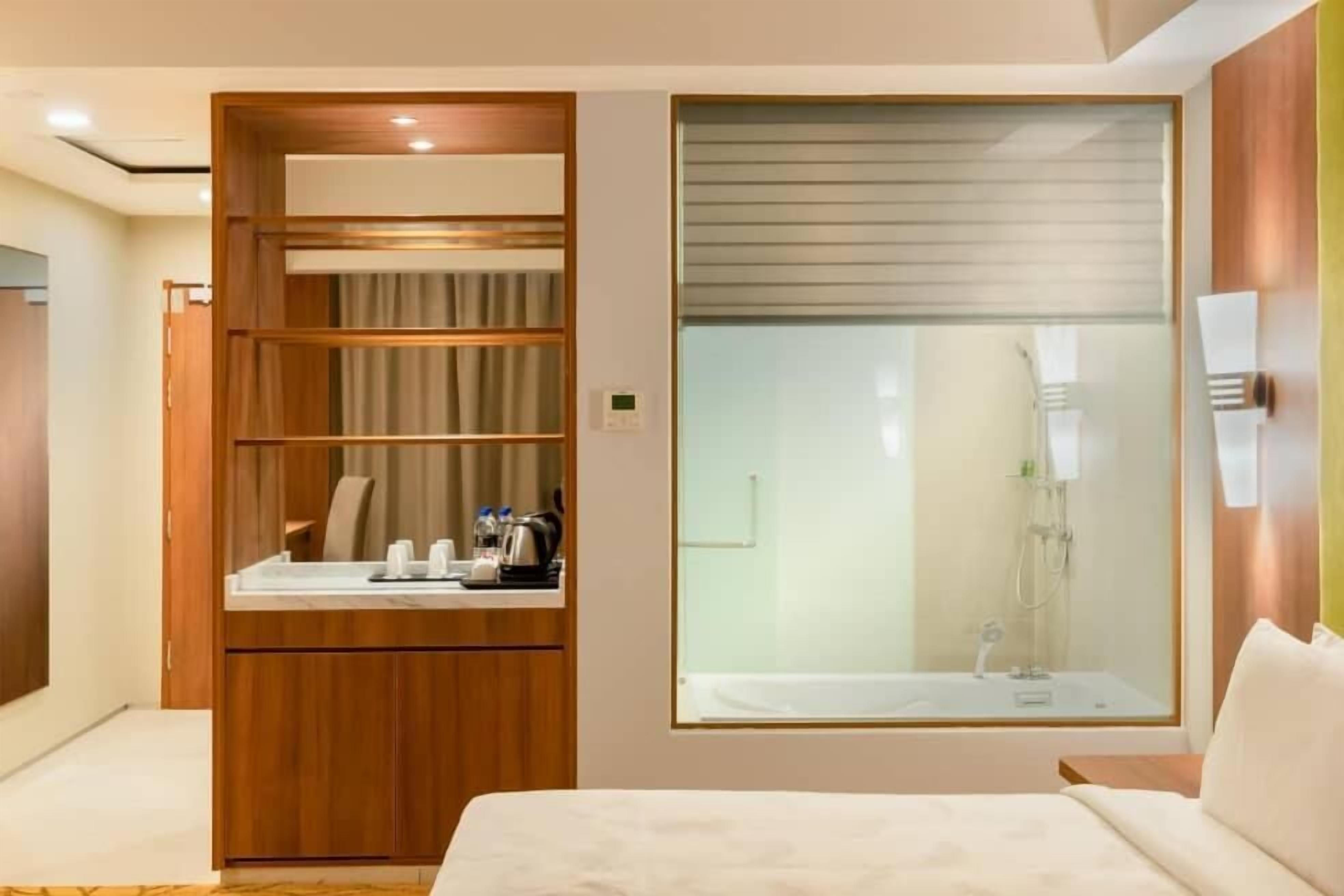 A hotel room with a bed, a wooden wardrobe, a bathroom with a glass shower enclosure, and a small table with toiletries.