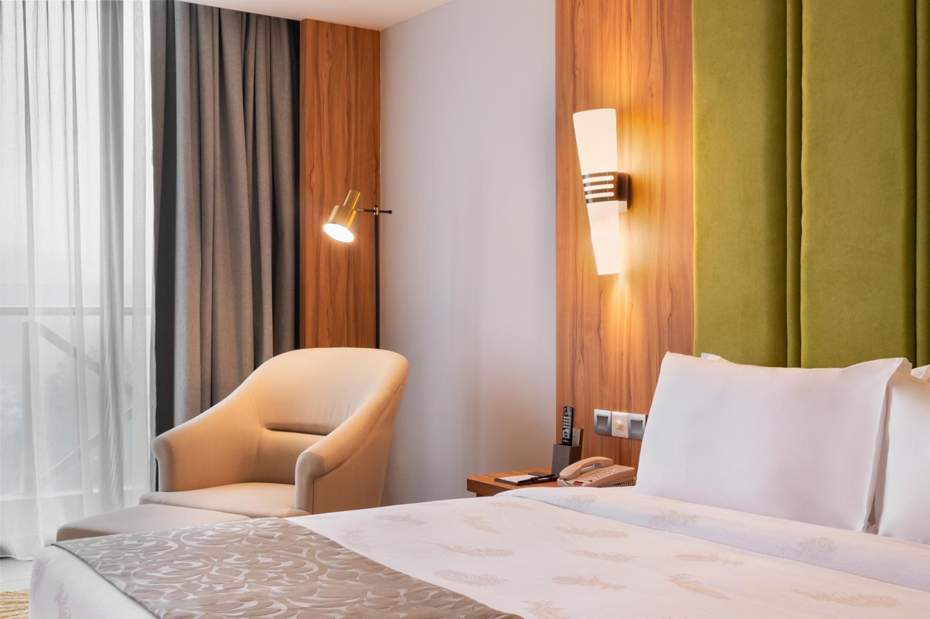Una habitación de hotel con una cama, una silla, una mesita de noche con teléfono, y una lámpara fija en la pared.