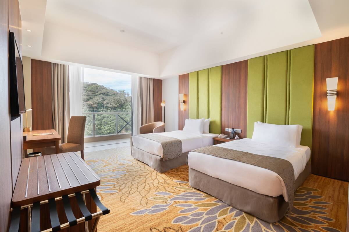 A hotel room with two beds, a desk, and a view of the outdoors.
