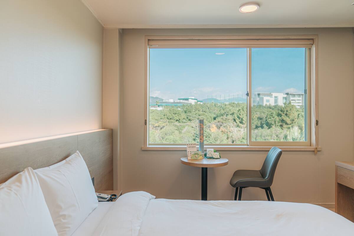 Habitación de hotel con cama, una silla, una mesita y ventana con vistas a zonas verdes y edificios.