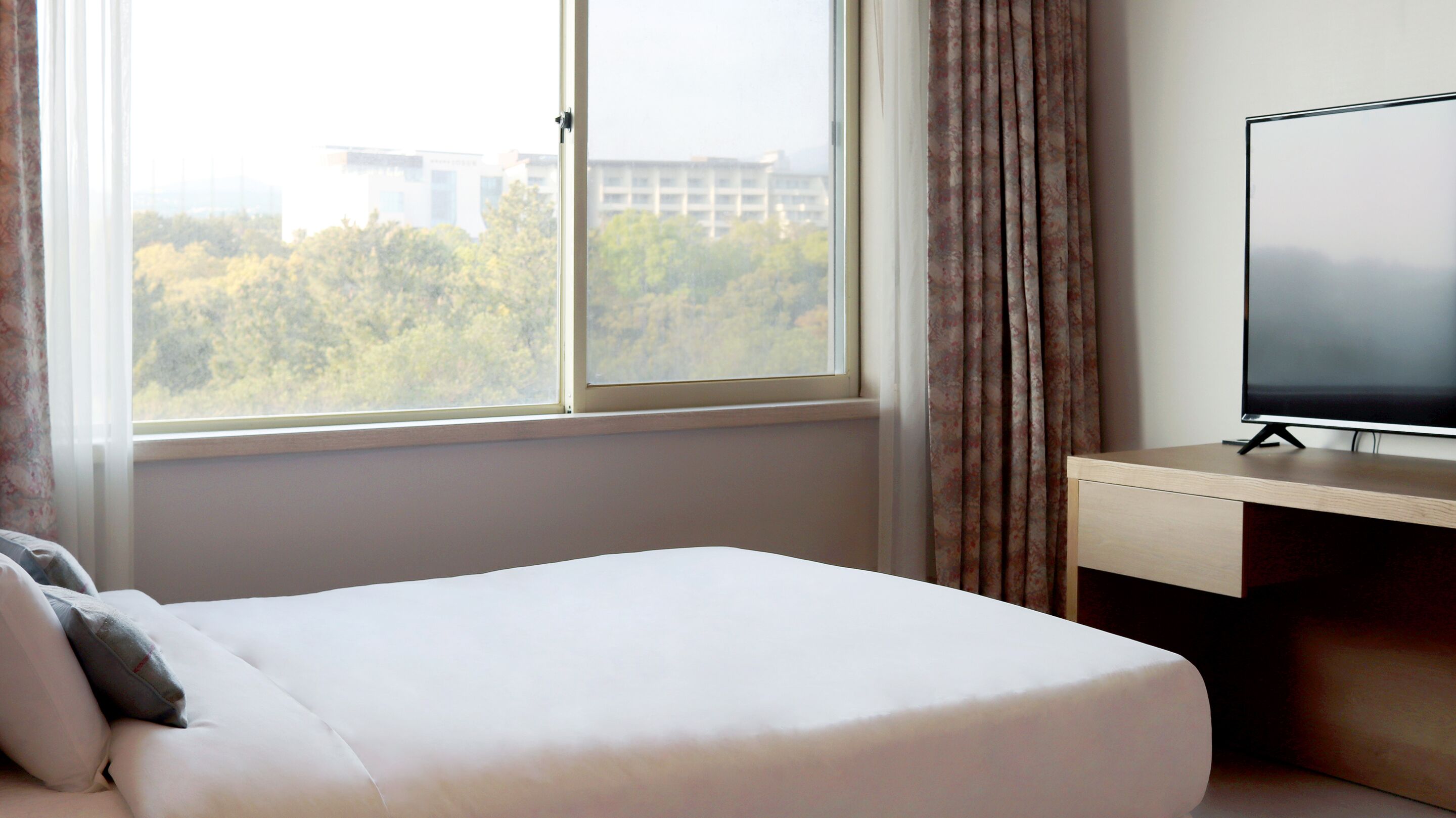 A hotel room with a bed, a desk with a television, a window with curtains, and a view of buildings and trees outside.