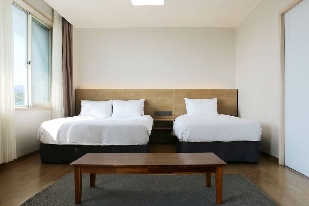 Una habitación de hotel con dos camas, una mesa de madera y un amplio ventanal con cortinas.