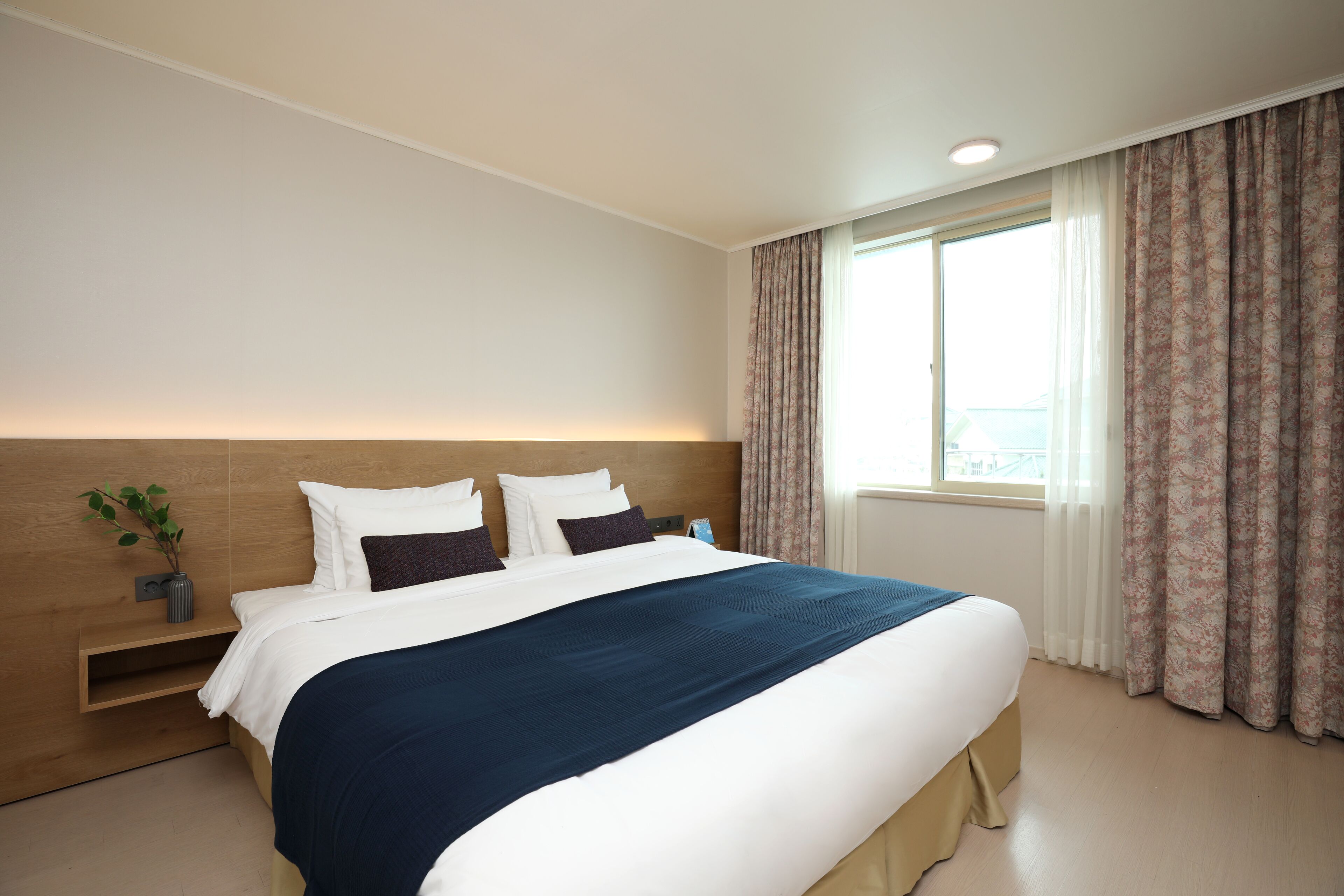 A hotel room with a large bed, a bedside table with a plant, a window with curtains, and a view of the outside.