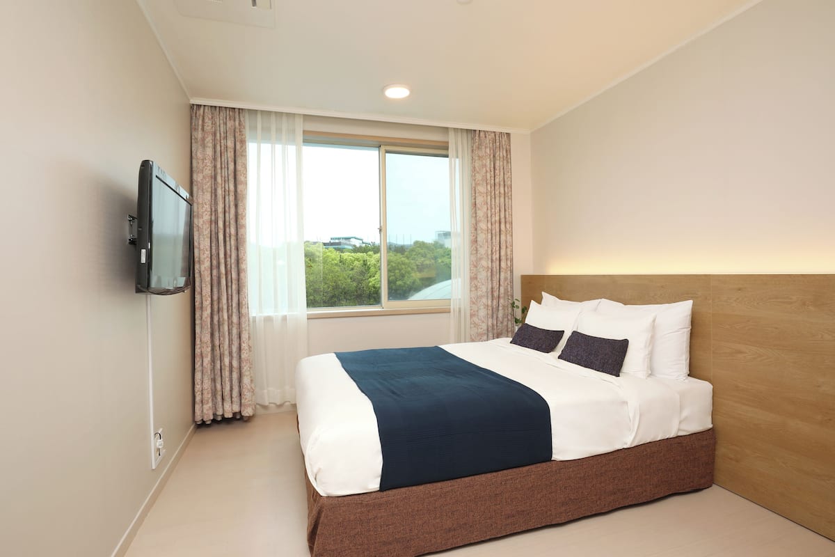 Una cama bien hecha con cabecera de madera, un televisor montado en la pared y una ventana con cortinas.