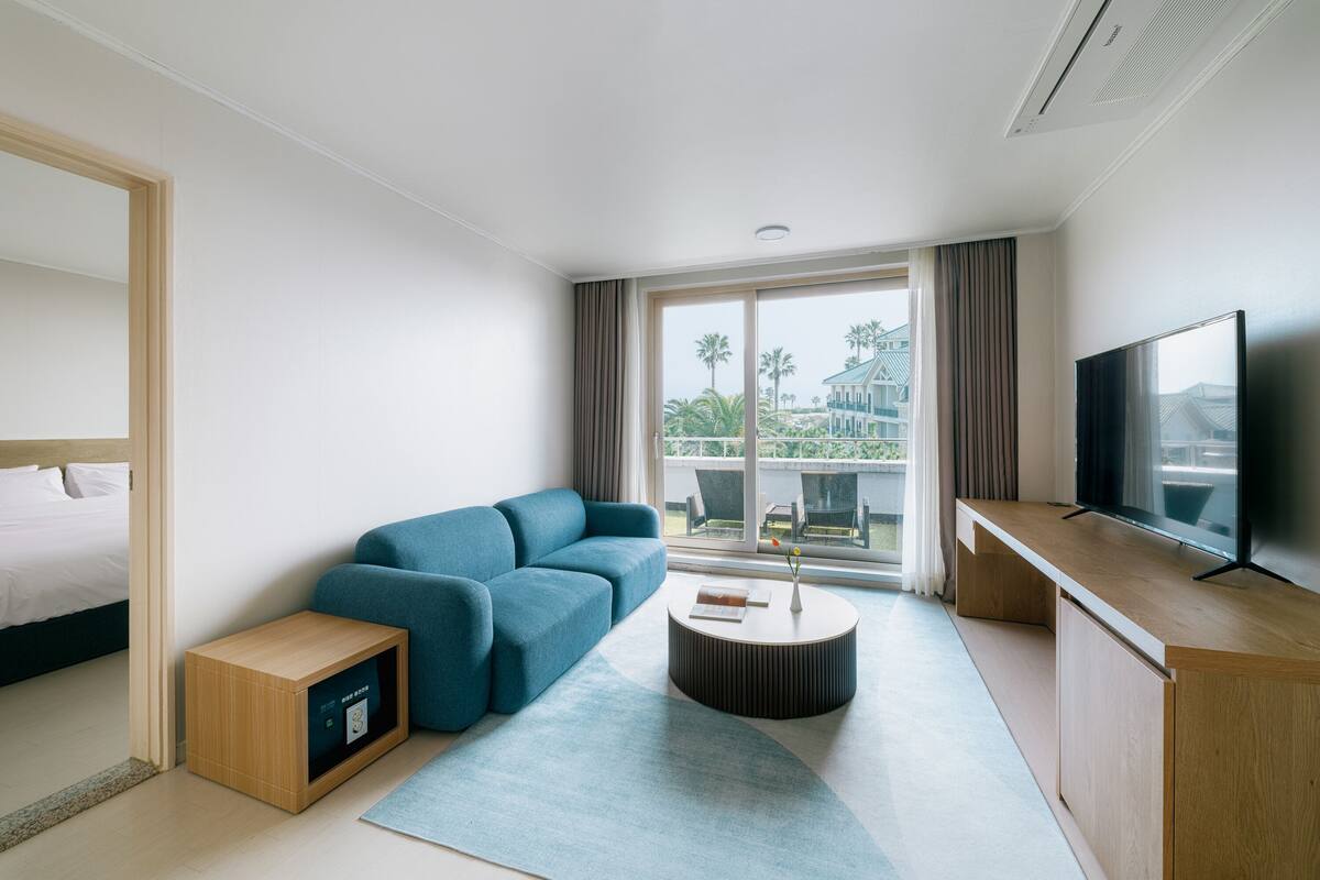 Una habitación de hotel moderna con cama, sofá azul, mesita de madera, mesa de centro redonda, televisor y ventana grande con vistas a palmeras y un edificio.