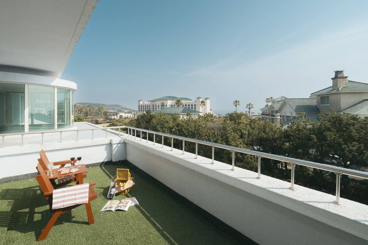 Una terraza en la azotea con baranda blanca, una mesa de madera con sillas y vista a un edificio grande en la distancia.
