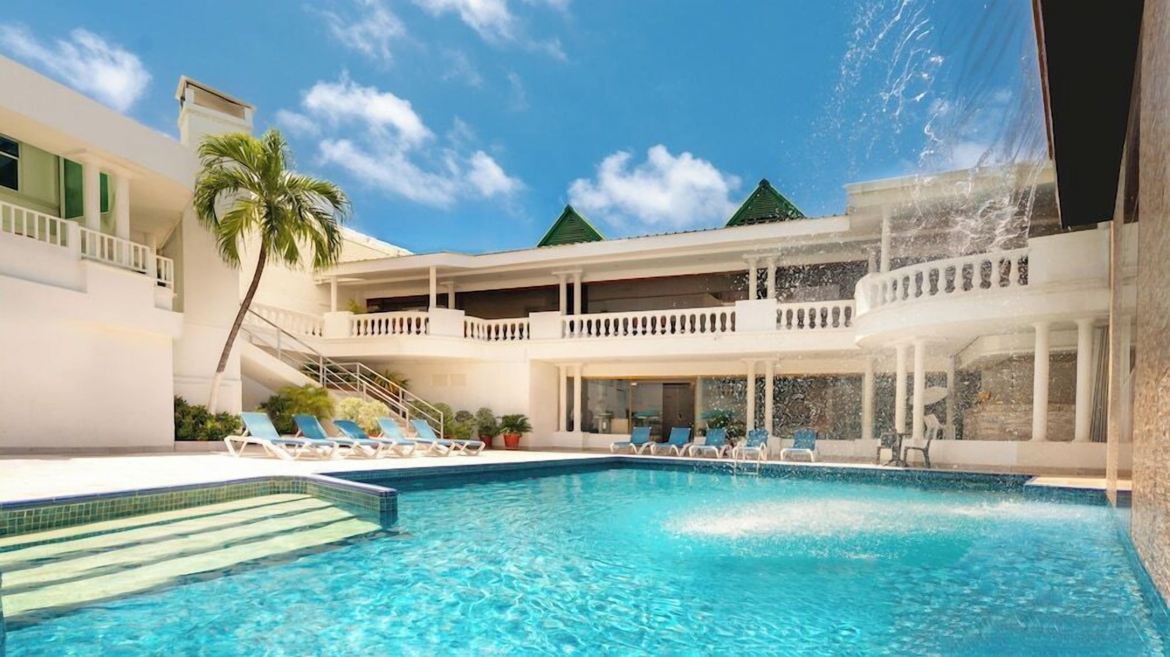 A swimming pool with lounge chairs and a palm tree, surrounded by a building with balconies and a clear sky.