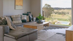 Ein modernes Wohnzimmer mit einer grauen Sofagarnitur, einem großen Fenster mit Blick auf eine ländliche Landschaft und einem Holztisch.