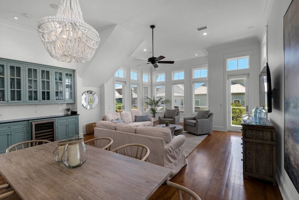 A spacious living area with a high ceiling, a chandelier, and a ceiling fan. There is a dining table with chairs, a sofa, and a TV mounted on the wall.